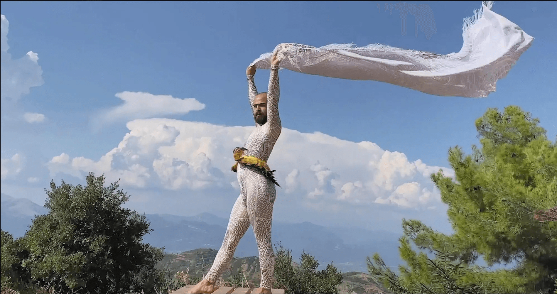 A performer in a white sequined bodysuit holds a length of white fabric billowing overhead against a bright sky and distant hills.