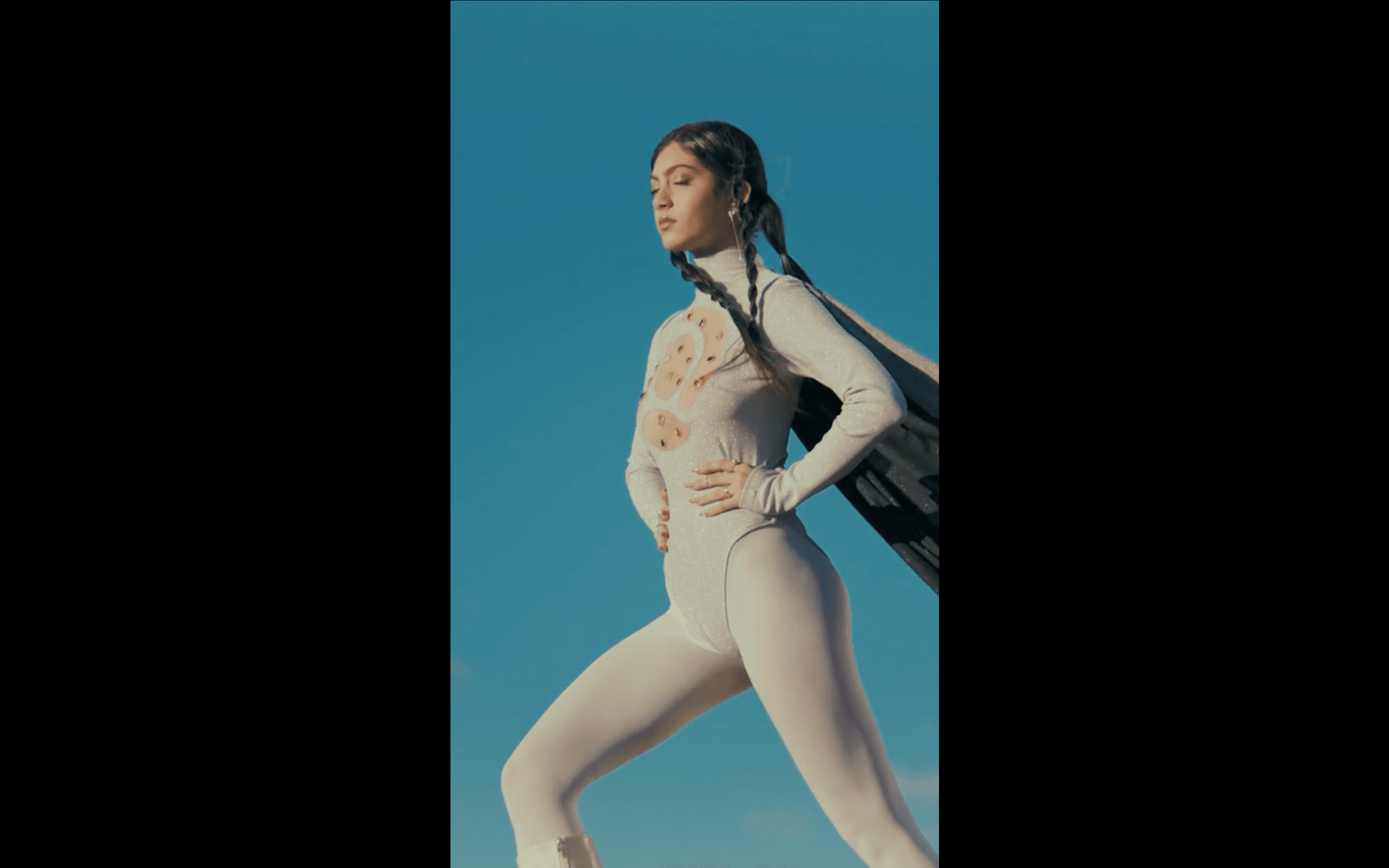 Shot from below against a vivid blue sky, a young Latin American woman in a white patterned bodysuit and long braids stands in a wide-legged pose with her hand on her hip. The low angle and open sky give the image a bold, empowered quality.