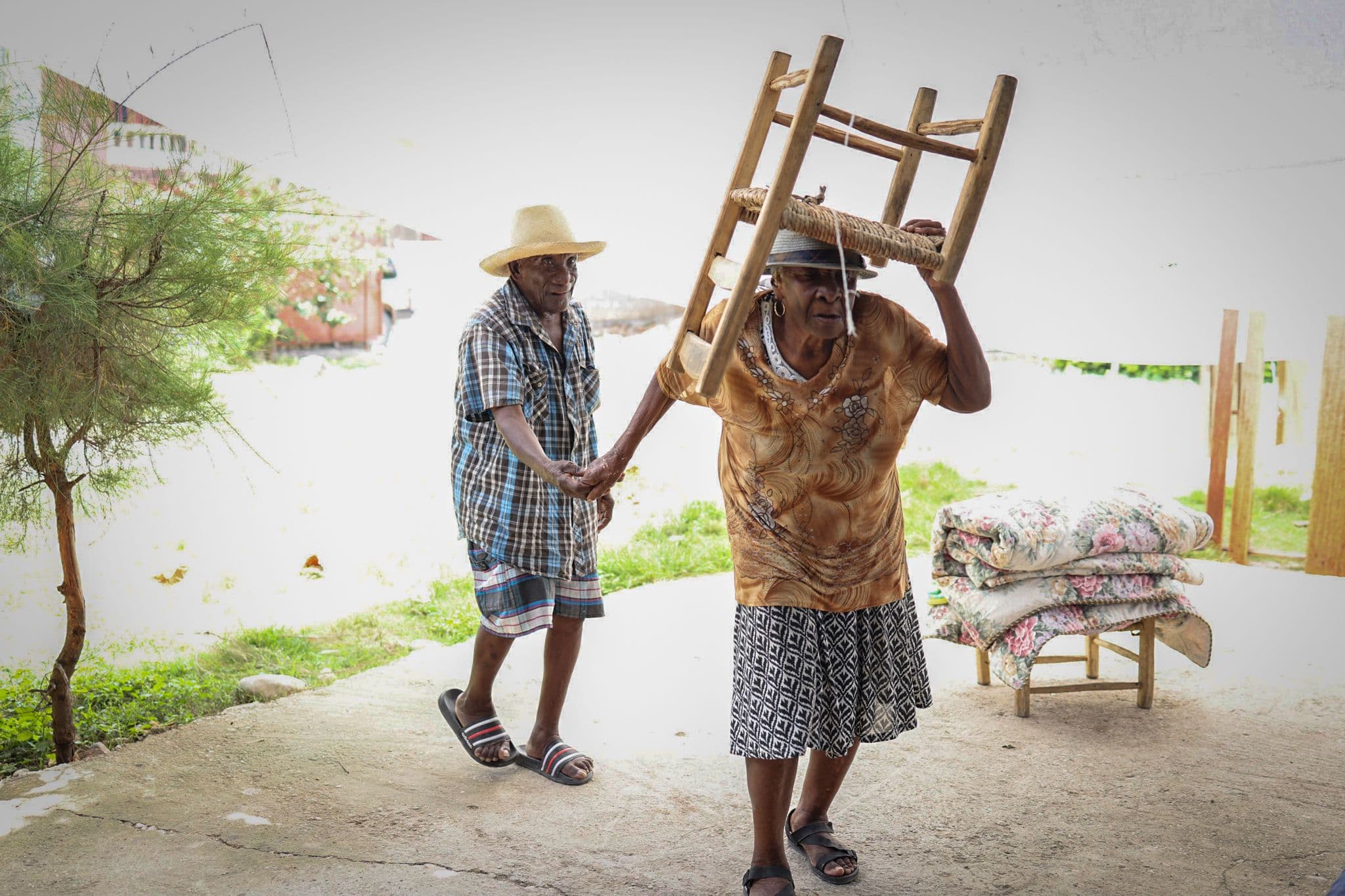 Two people carry a wooden chair and bundled bedding along a path, relocating belongings after a disaster.