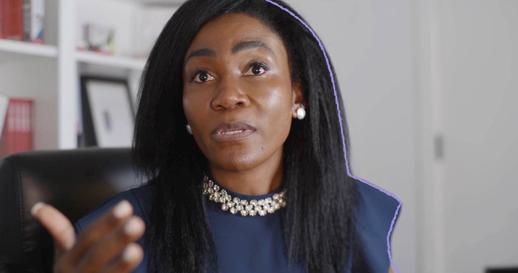 A woman with a beaded necklace speaks and gestures mid-interview in an office, traced with a glowing lavender outline.