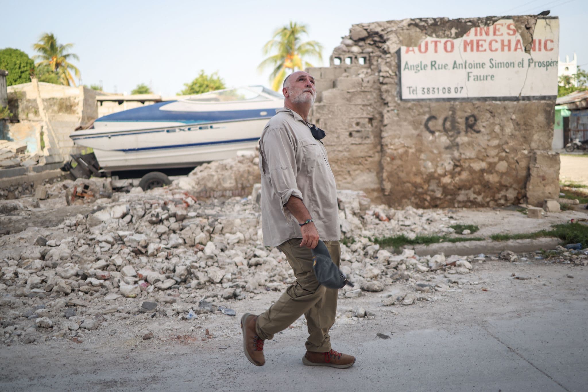 A man walks past a pile of rubble and a battered fishing boat beside a damaged building marked 'Auto Mechanic,' surveying earthquake damage.