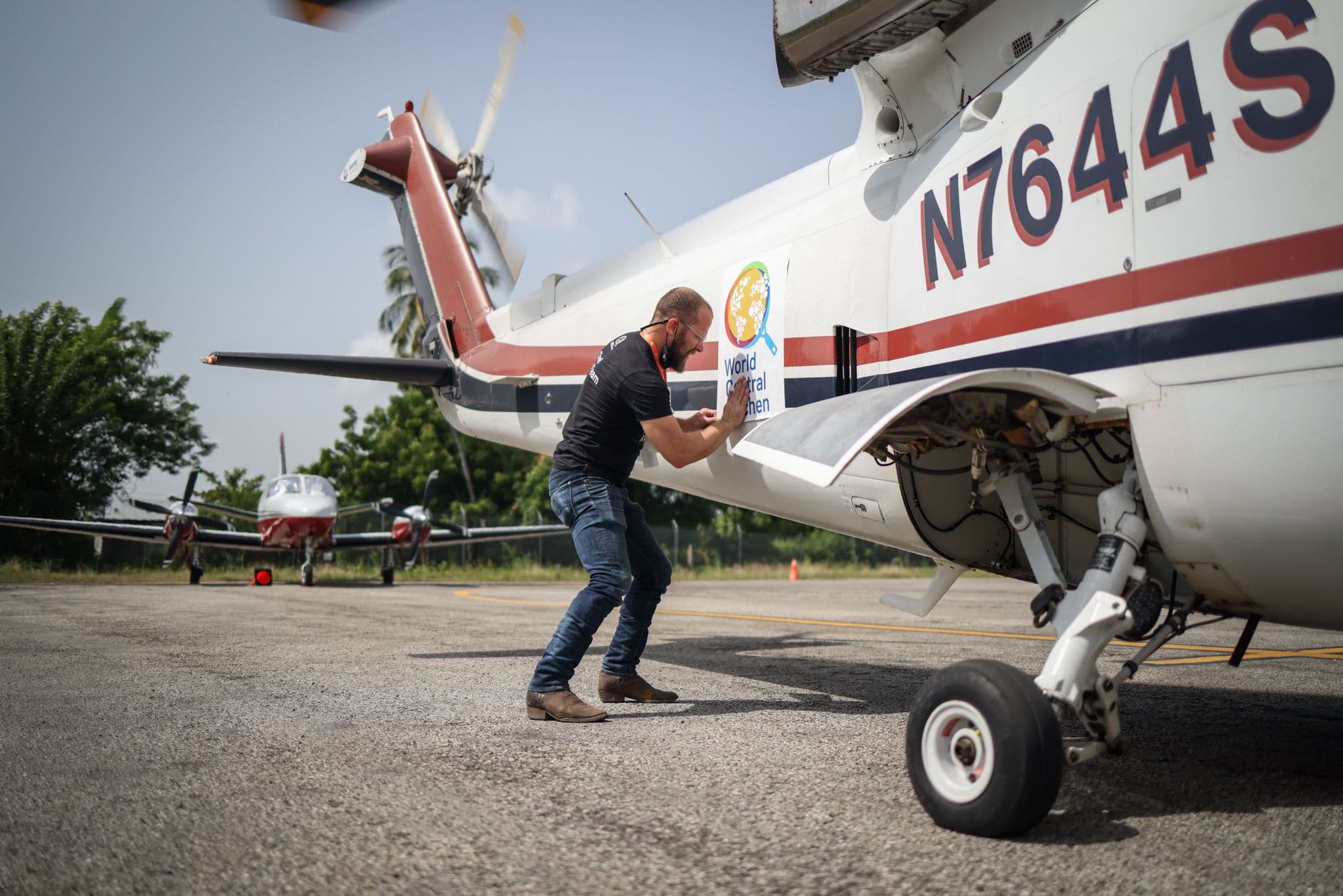 A man loads a box of supplies into the cargo hold of a small propeller plane on an airport tarmac.
