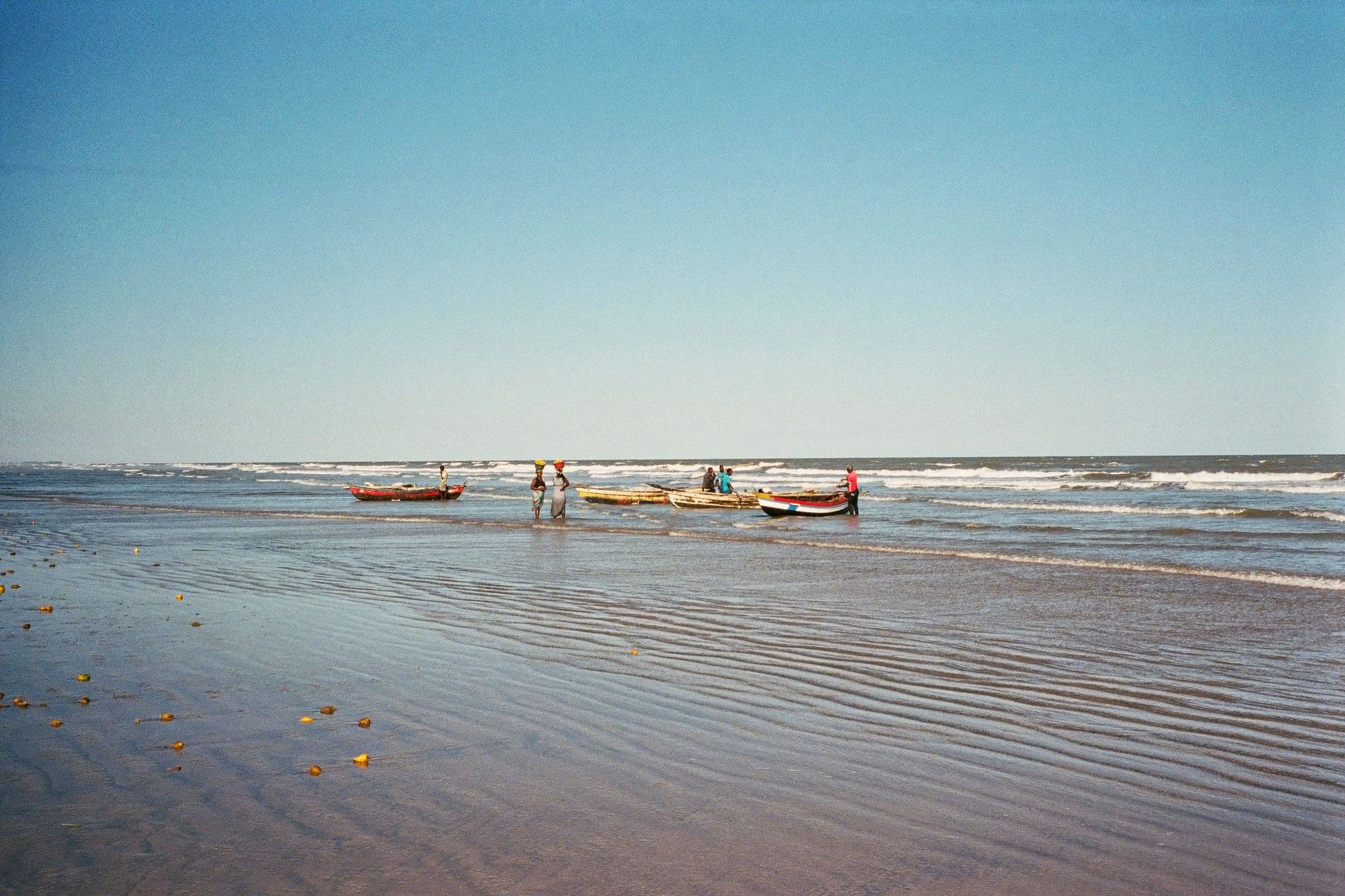 A group of people and several wooden canoes gather at the water's edge on a wide sandy beach.