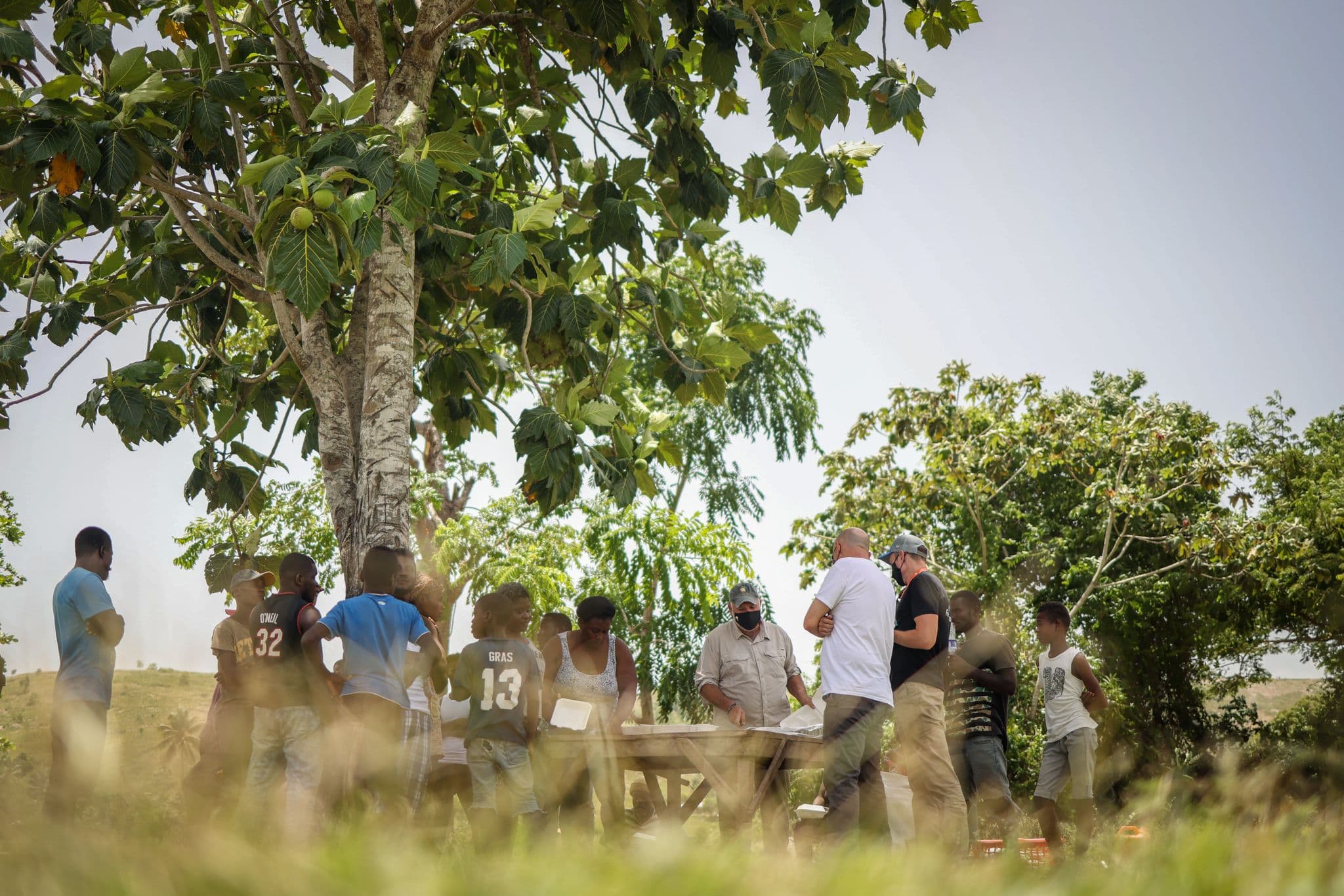 A group of people gathers in the shade of a large tree in a rural field, standing around a wooden table.