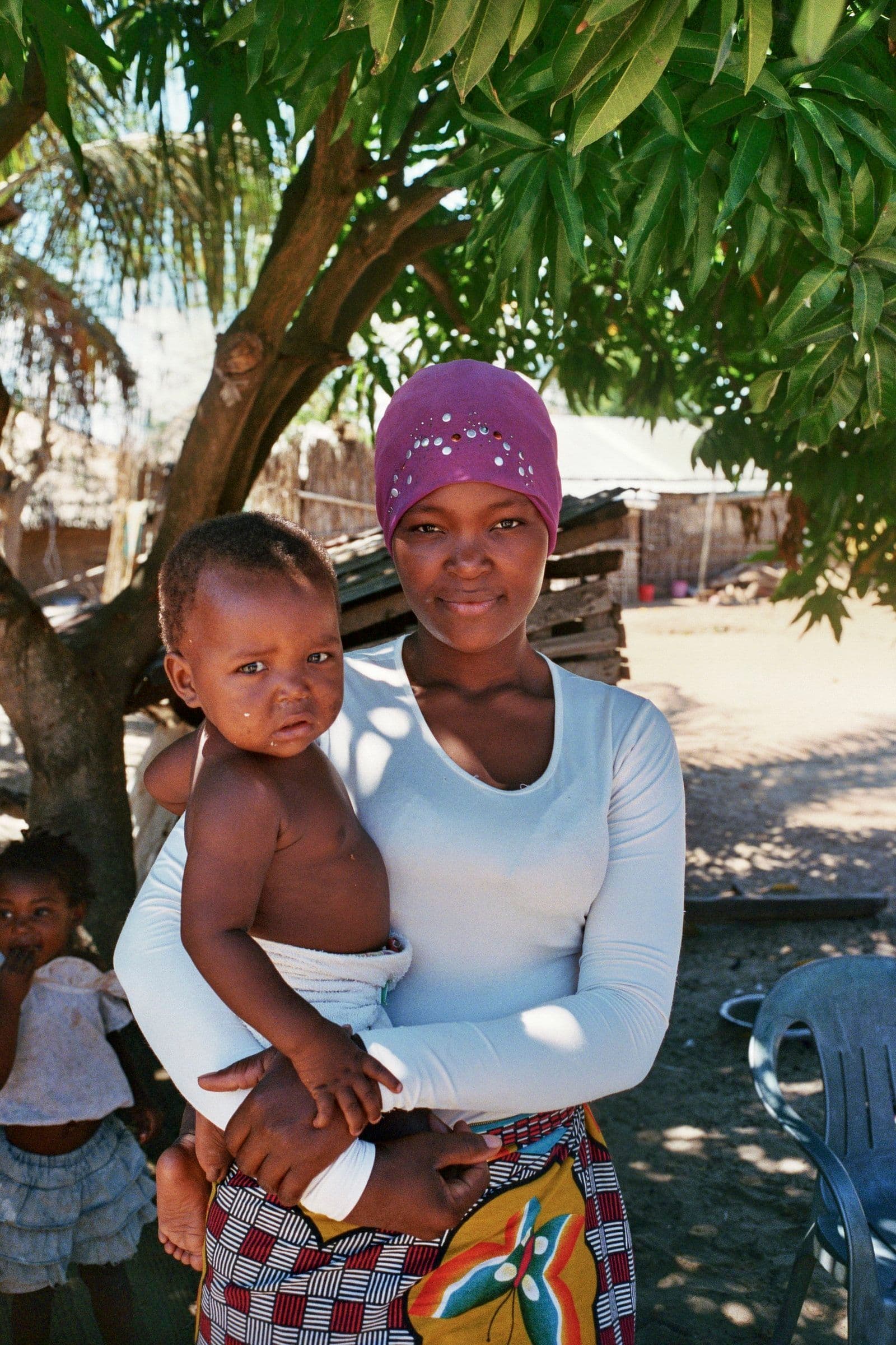 A woman in a purple headwrap holds a young child under a shade tree, with more children visible nearby.