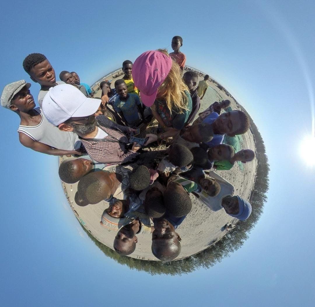 A distorted 360-degree 'tiny planet' photo showing a circle of children and adults gathered together outdoors under a bright sky.