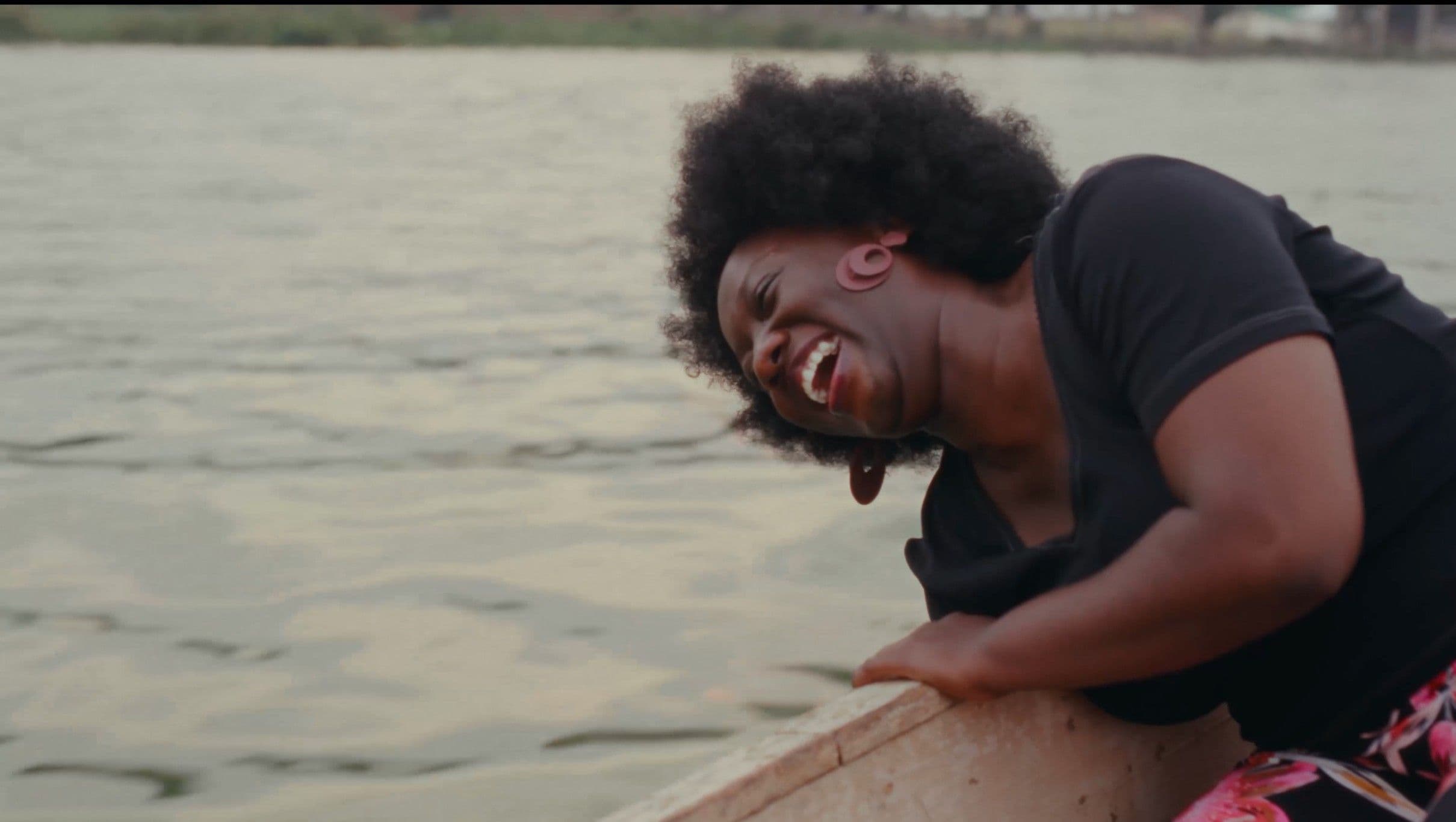 A Black woman leans over the edge of a wooden boat on a wide river, laughing openly with raw, uninhibited joy. The warm, hazy light on the water behind her adds an intimate, documentary quality to the moment.
