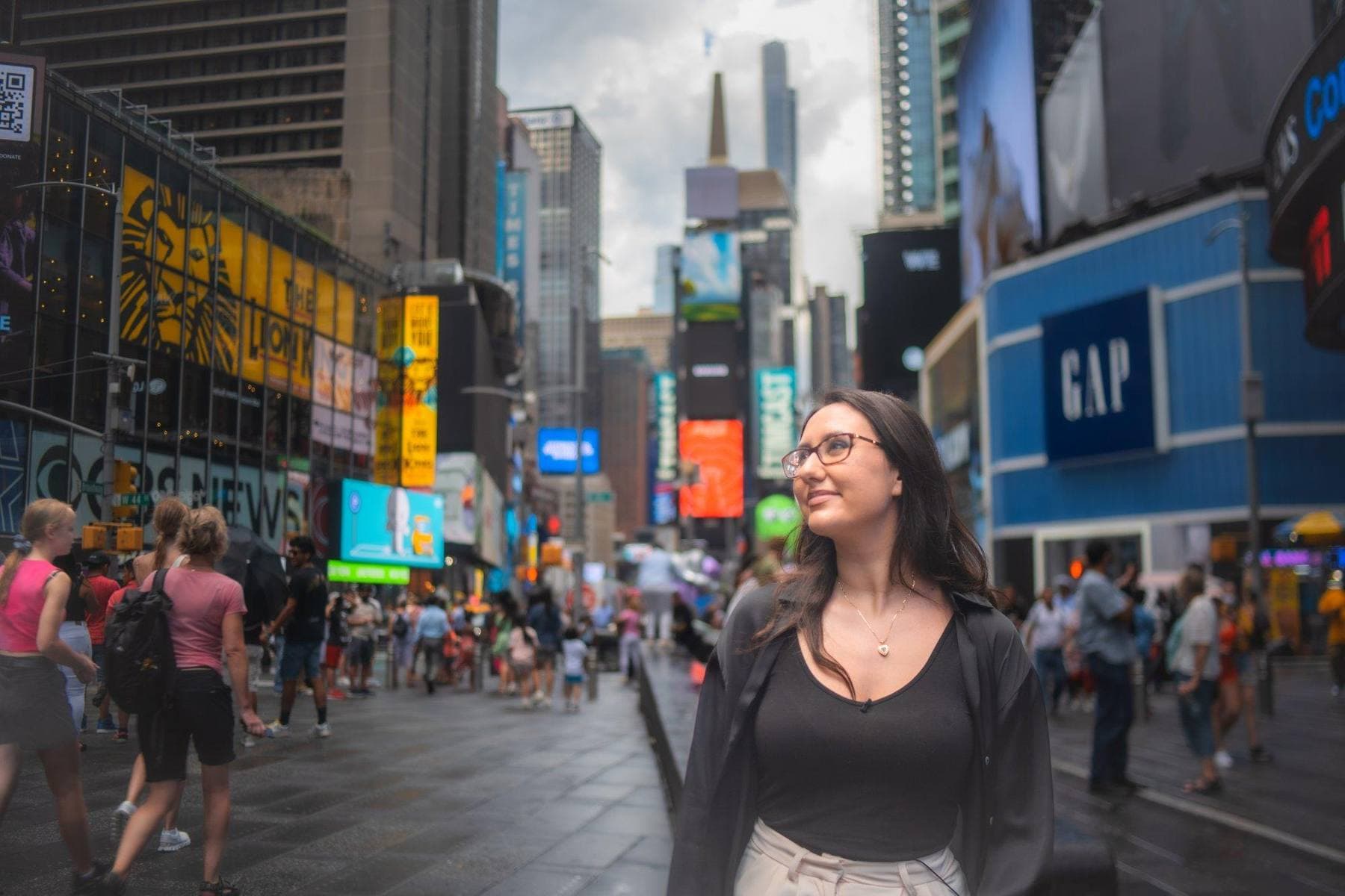 A young woman stands in a crowded Times Square at dusk, looking up past the billboards and city lights.