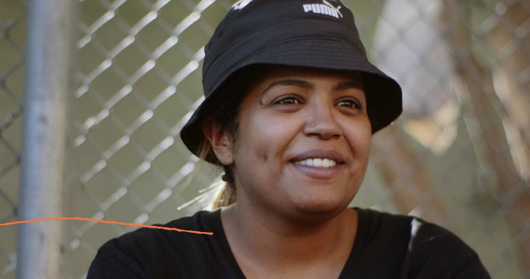 A woman in a black Puma bucket hat smiles warmly in front of a chain-link fence, a thin orange line tracing across the frame.