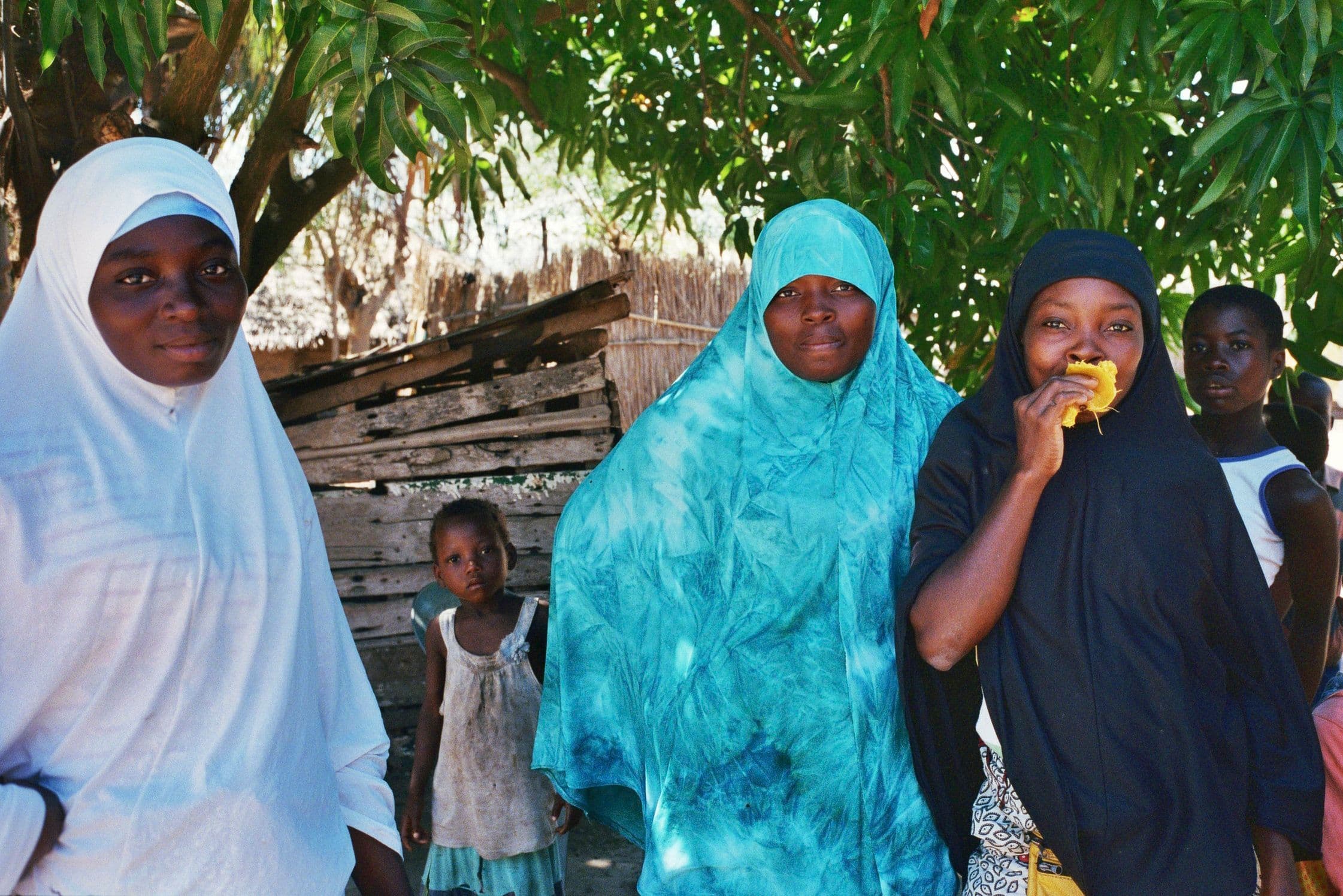 Three women stand together under shade trees, one in a white headscarf, one in teal tie-dye, and one in black, with children in the background.