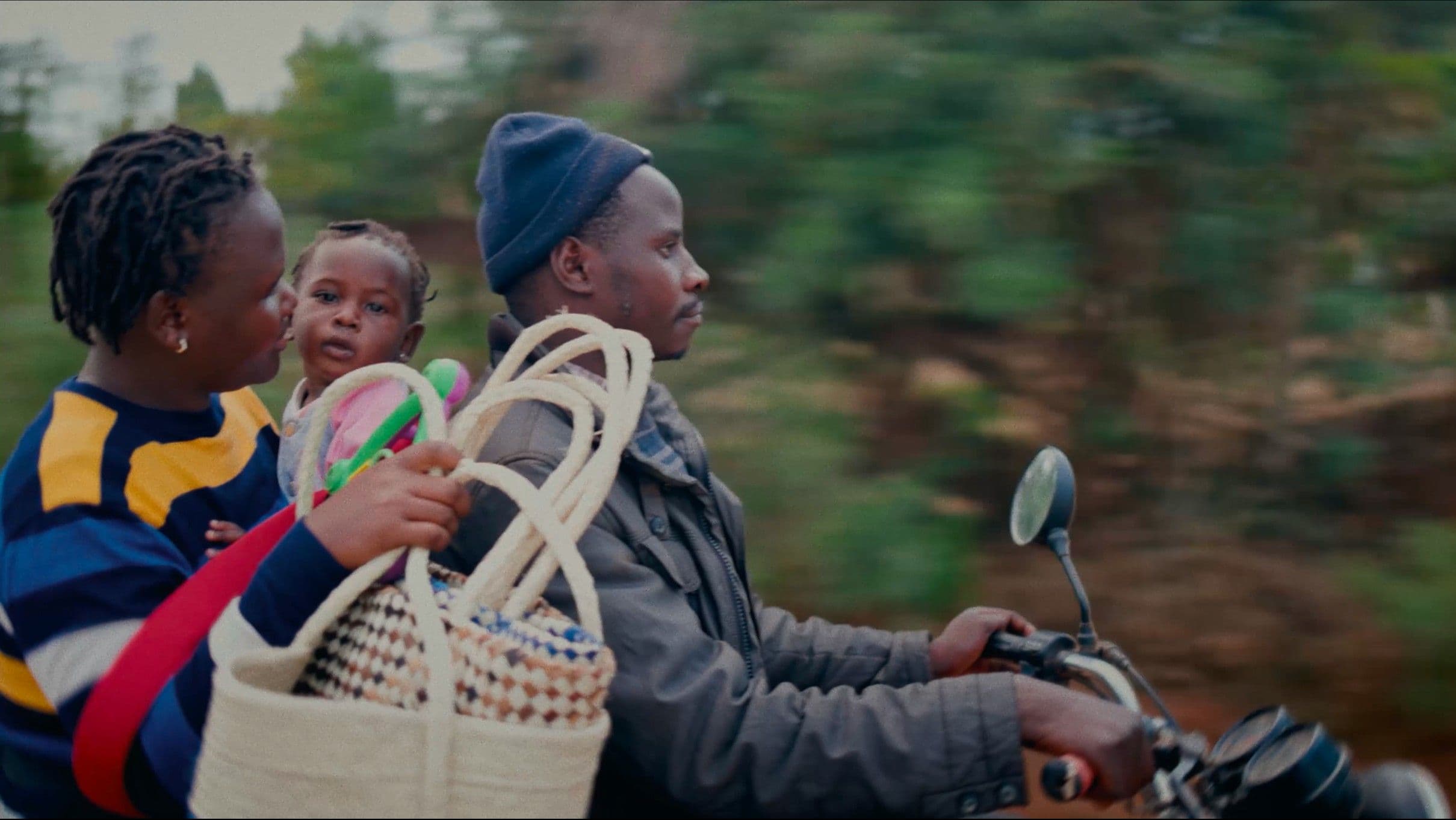 A man rides a motorbike through a lush green landscape while a woman seated behind him holds a young baby and a woven basket. The motion-blurred background gives the cinematic still a sense of journey and everyday resilience.