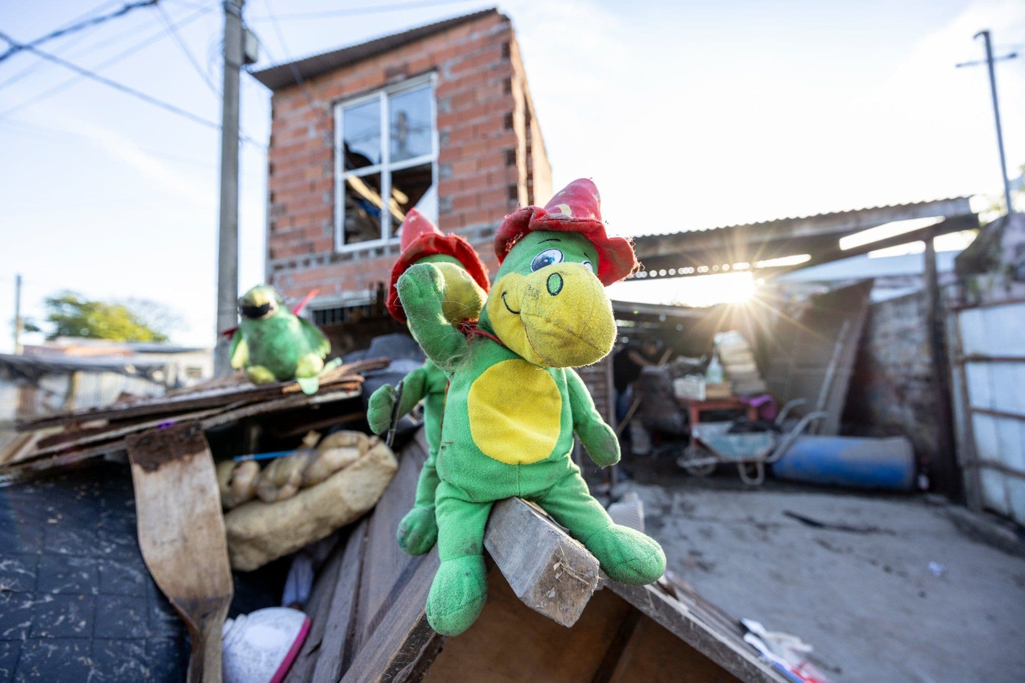 A green stuffed toy dragon sits atop a boat surrounded by flood debris, with a damaged brick building in the background.