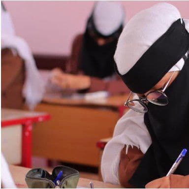 A photo of a girl in a white headscarf and glasses writing at her desk in a classroom, with classmates seated behind her.