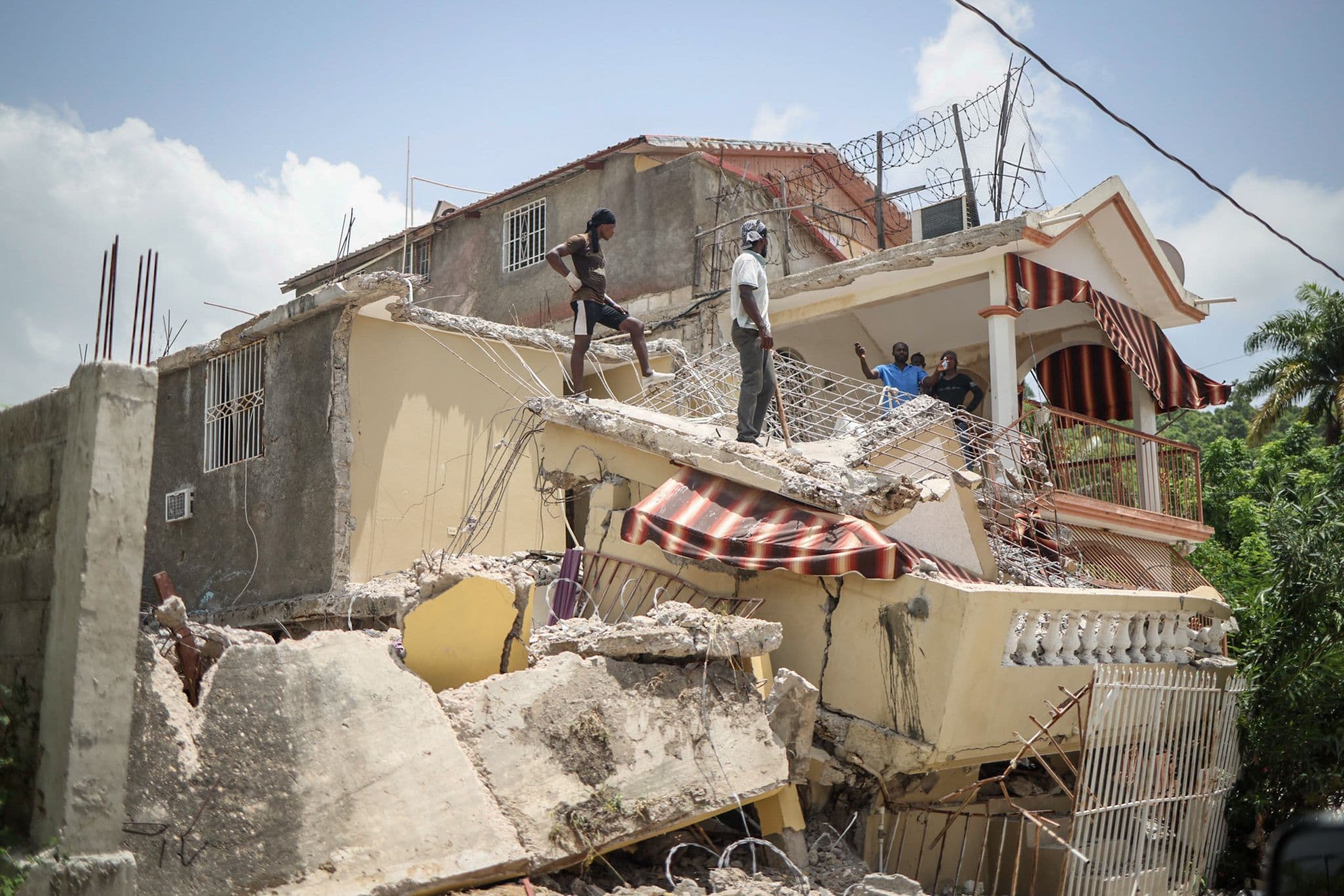 People stand atop the rubble of a collapsed multi-story building after an earthquake, its balcony and awning crushed beneath the debris.